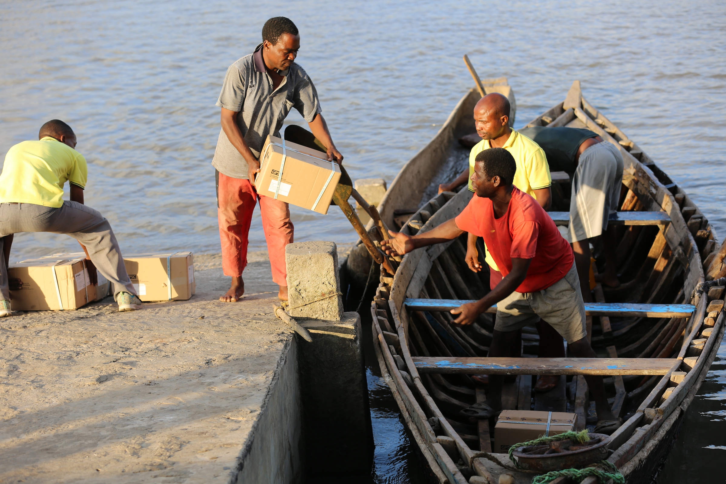 Men load cardboard boxes embossed with the Coca-Cola logo from a dock into a wooden boat on the water.
