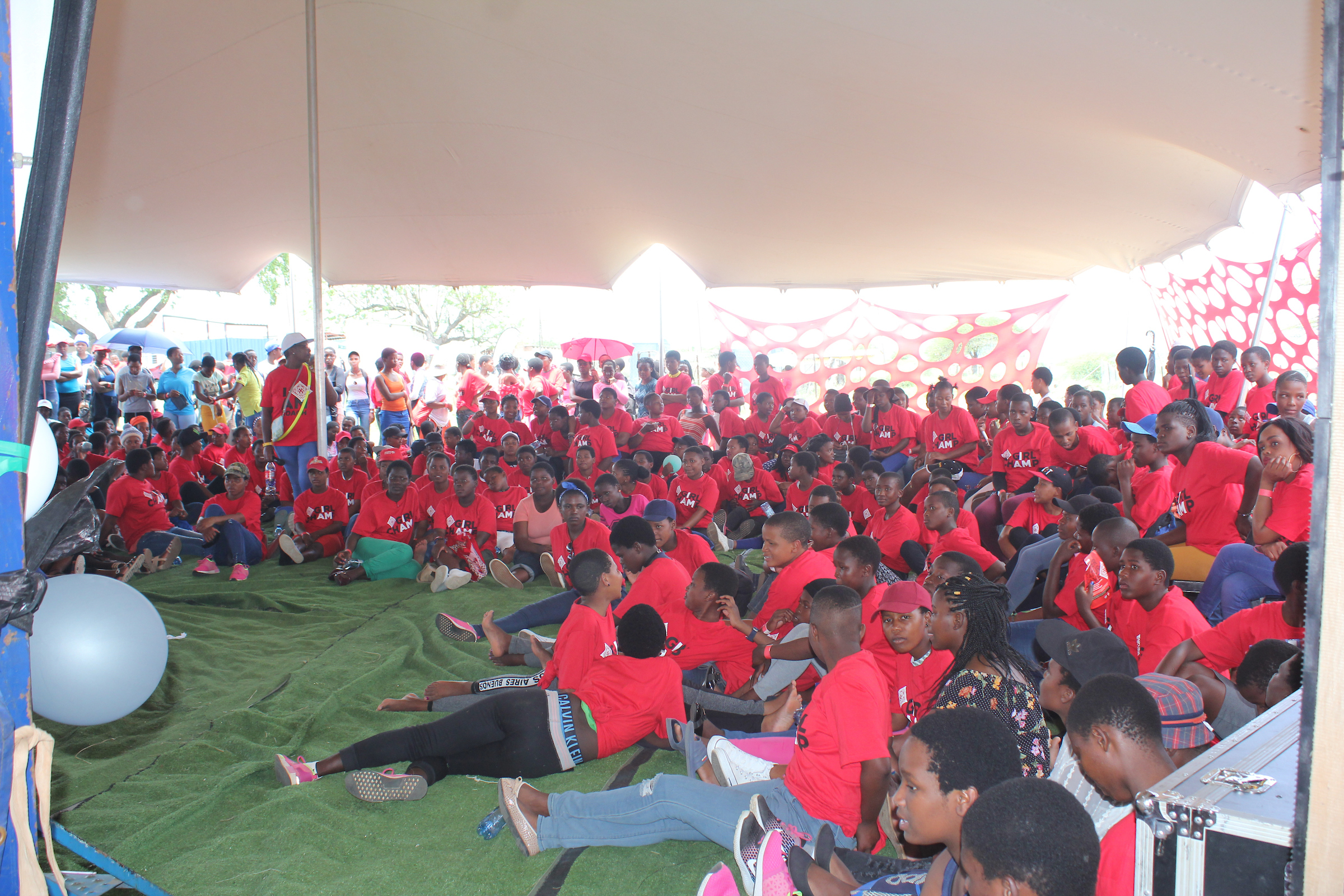 A large group of people, mostly wearing red shirts, including many young girls, sit under a tent on a green carpet at an outdoor event in Eswatini.
