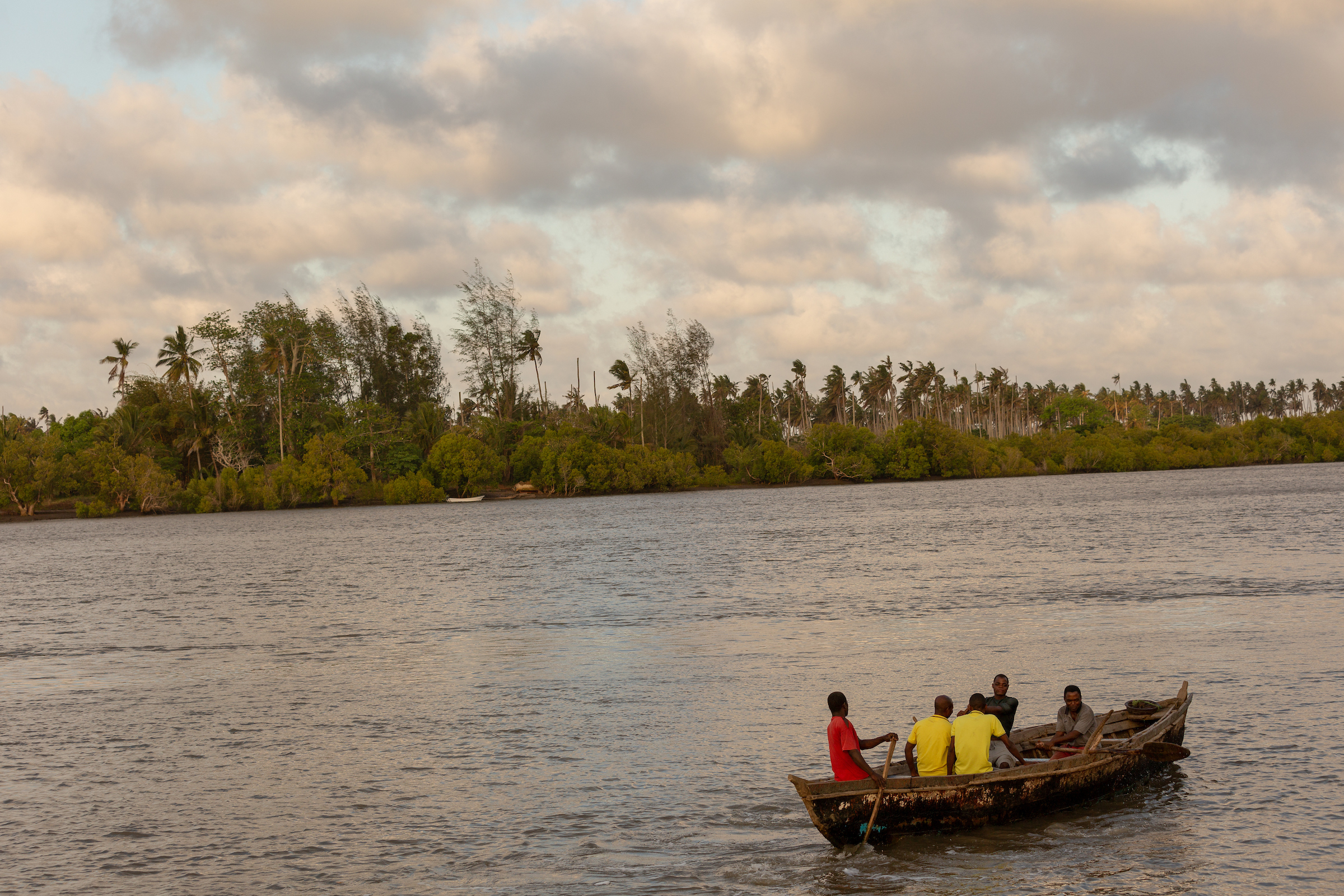 A group of people in a small wooden boat on a calm river navigates through Mozambique's lush, tree-lined shores under cloudy skies, exploring nature's tapestry.