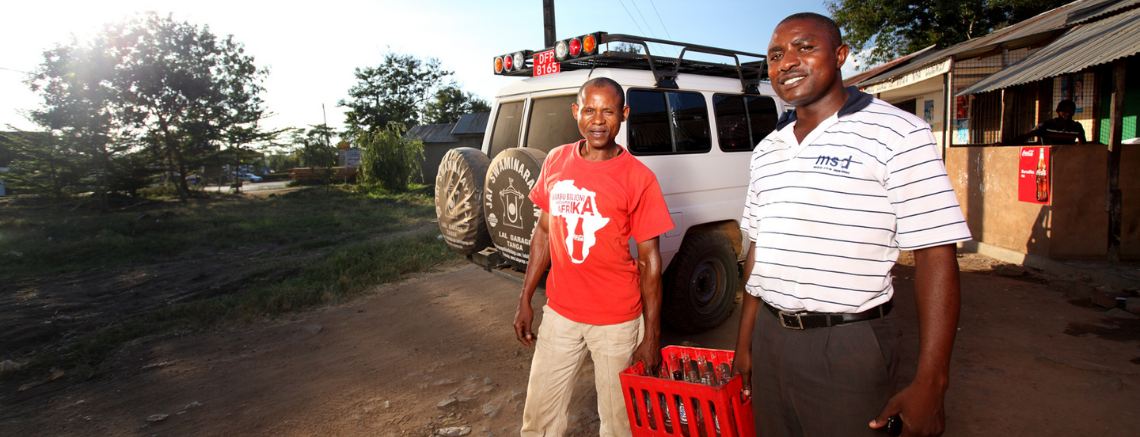 Two men stand outdoors, one in a red shirt holding a crate, and the other in a striped shirt. A vehicle and building loom in the background, hinting at their roles within a global health partnership that thrives on trust.