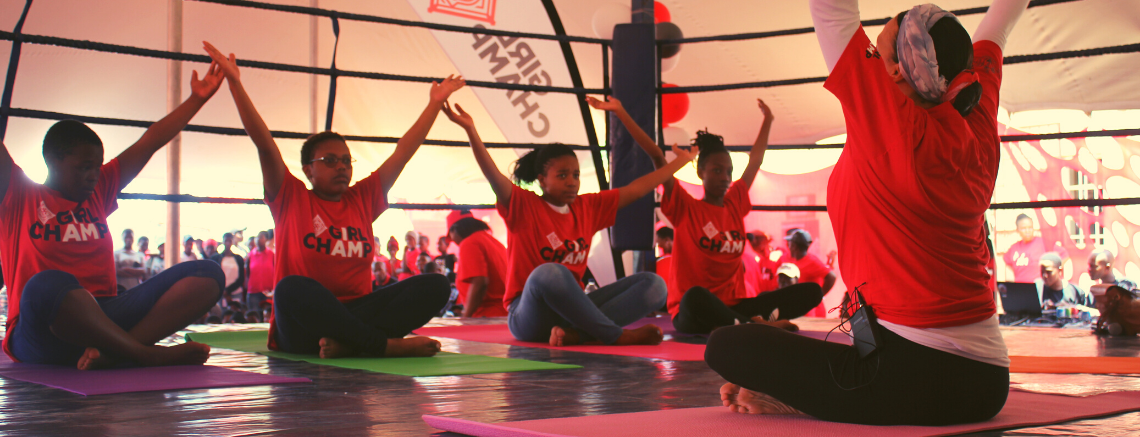 A group of adolescent girls in red shirts sit cross-legged on mats, raising their arms, in a Girl Champ tent-like setting.